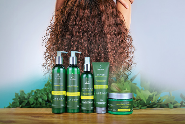 Produtos da linha Curl & Wave da Grandha expostos em frente a um cabelo cacheado, destacando os itens de cuidados como shampoo, condicionador, leave-in e máscara.