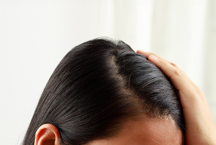 Close-up do couro cabeludo de uma mulher com cabelos pretos e lisos, enquanto ela afasta o cabelo com a mão, destacando a raiz saudável.