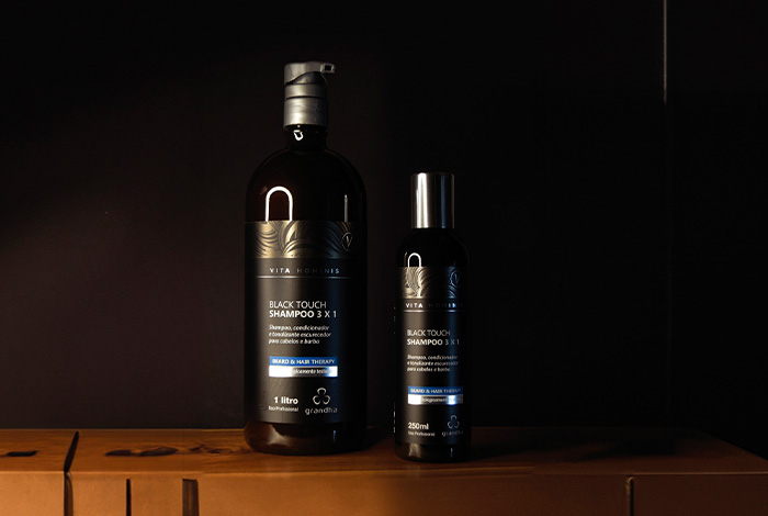 Dois frascos do shampoo Black Touch 3x1 da Grandha, em tamanhos diferentes, sobre uma superfície de madeira com fundo preto elegante.