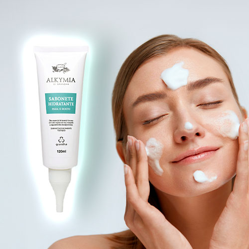 Mulher aplicando espuma de sabonete no rosto com expressão relaxada ao lado de um sabonete facial hidratante da linha Alkymia da Grandha.