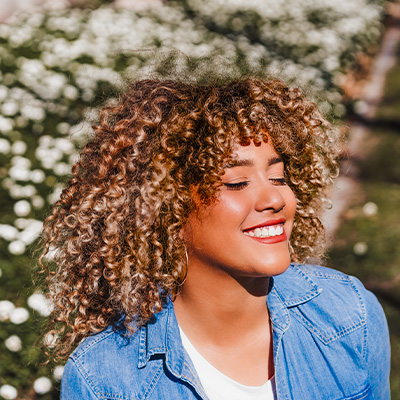 Mulher de cabelos cacheados, com luzes, sorrindo de olhos fechados sob um fundo desfocado com petalas brancas.