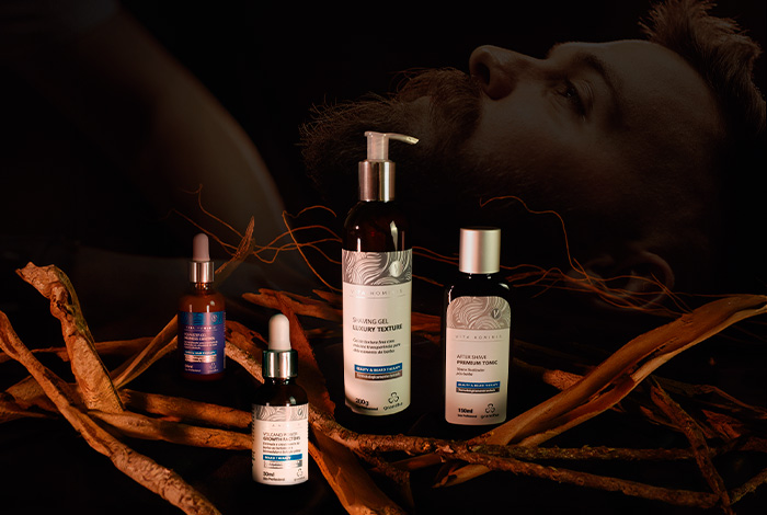 Kit de cuidados com a barba da linha Vita Hominis da Grandha, com frascos sobre raízes e homem barbudo ao fundo em clima escuro e elegante.