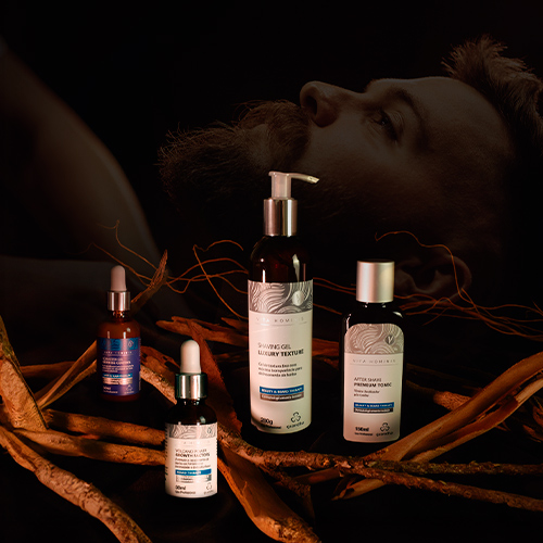 Kit de cuidados com a barba da linha Vita Hominis da Grandha, com frascos sobre raízes e homem barbudo ao fundo em clima escuro e elegante.