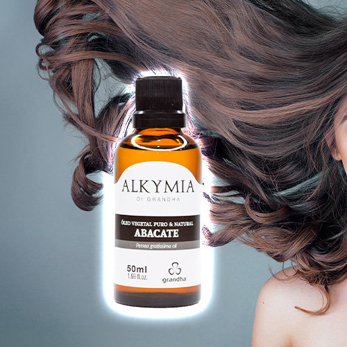 Pessoa com cabelos longos soltos ao vento ao lado de um frasco de óleo vegetal de abacate Alkymia di Grandha, usado para cuidados capilares.