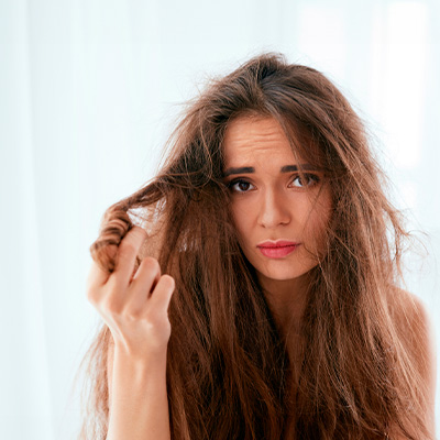 Mulher com expressão preocupada observa os próprios cabelos ressecados e com frizz, segurando uma mecha com a mão.