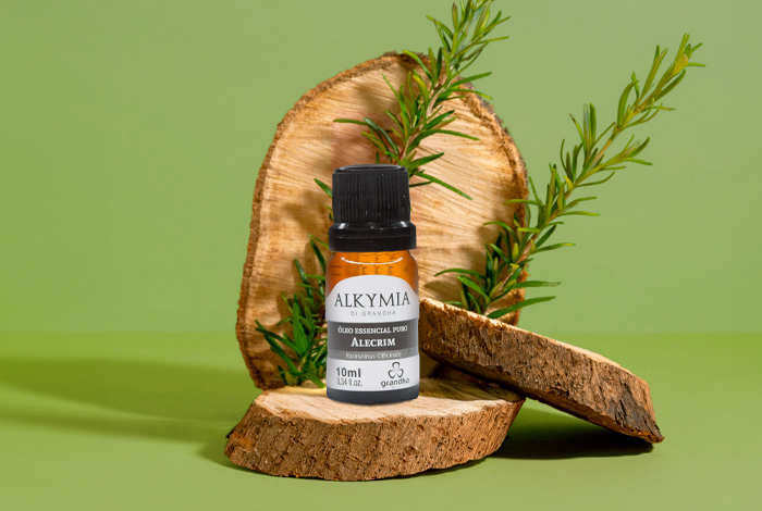 Frasco de óleo essencial de alecrim Alkymia sobre pedaços de madeira, com ramo de alecrim ao fundo e fundo verde natural.