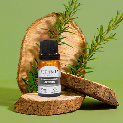 Frasco de óleo essencial de alecrim Alkymia sobre pedaços de madeira, com ramo de alecrim ao fundo e fundo verde natural.