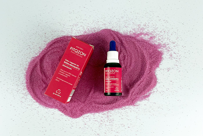 Frasco de óleo vegetal Fitozon Rosa Mosqueta da Grandha com caixa rosa, sobre fundo branco e areia rosa, em composição estética e natural.