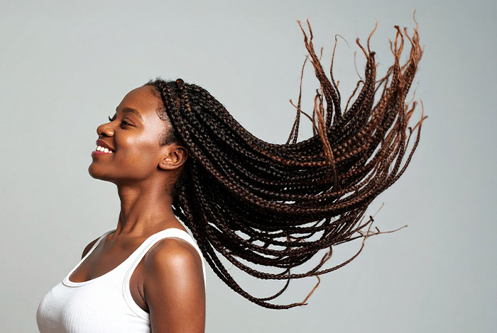 Mulher sorridente com tranças longas e esvoaçantes, usando regata branca, em fundo neutro.