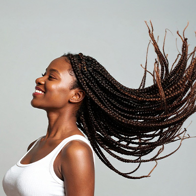 Mulher sorridente com tranças longas e esvoaçantes, usando regata branca, em fundo neutro.