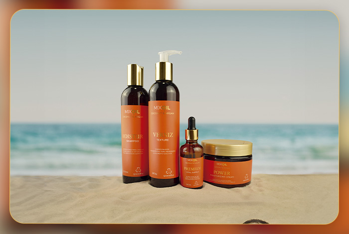 Conjunto de produtos Mix Oil Coconut & Argan sobre a areia, com o mar ao fundo, simbolizando hidratação intensa e cuidados capilares de verão.