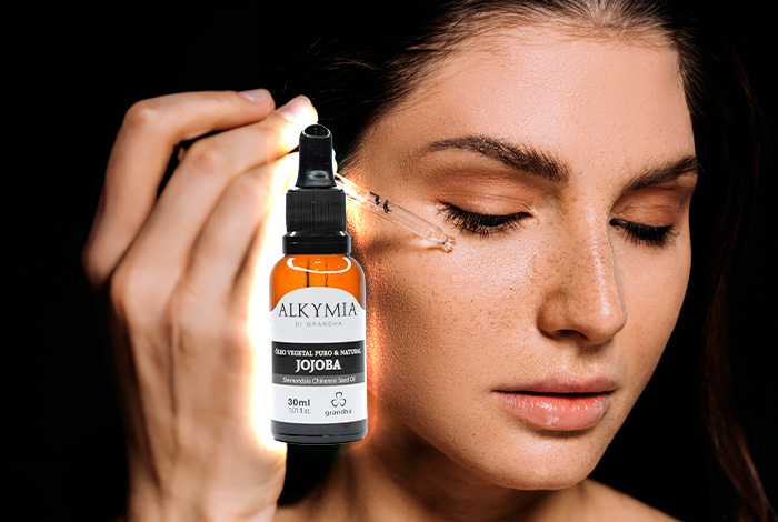 Mulher aplicando óleo de jojoba no rosto com conta-gotas, ao lado de frasco Alkymia di Grandha iluminado.