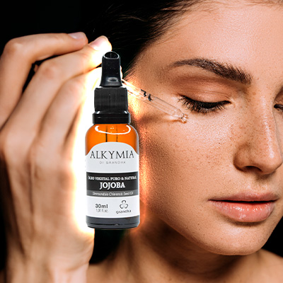 Mulher aplicando óleo de jojoba no rosto com conta-gotas, ao lado de frasco Alkymia di Grandha iluminado.