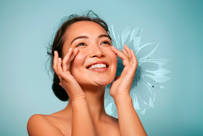 Óleo de girassol tira manchas da pele? Saiba mais. 1 Mulher sorridente toca suavemente o rosto com as mãos, pele iluminada, em fundo azul claro com flor clara ao lado do rosto.