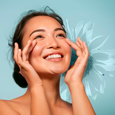 Mulher sorridente toca suavemente o rosto com as mãos, pele iluminada, em fundo azul claro com flor clara ao lado do rosto.