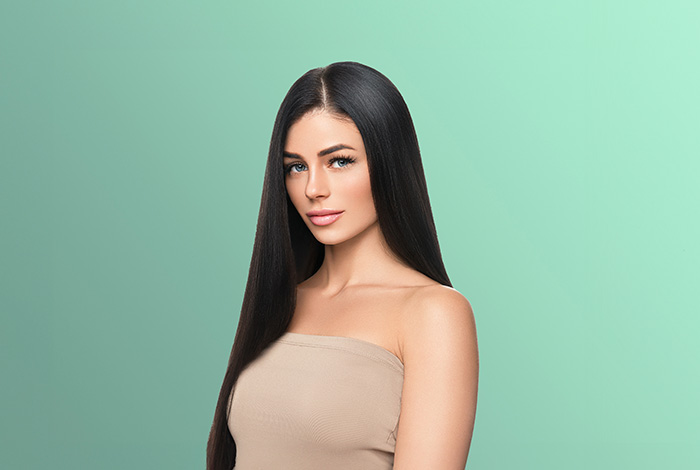 Alisamento capilar sem danos estruturais: é possível? 1 Mulher com cabelos longos, lisos e brilhantes posa de frente em fundo verde, destacando aparência saudável dos fios.