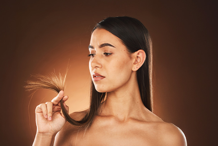 5 sinais de que seu cabelo precisa de reparação. 1 Mulher segurando a ponta dos cabelos e observando os fios ressecados e danificados, com cabelos longos e lisos sobre fundo em tons quentes.